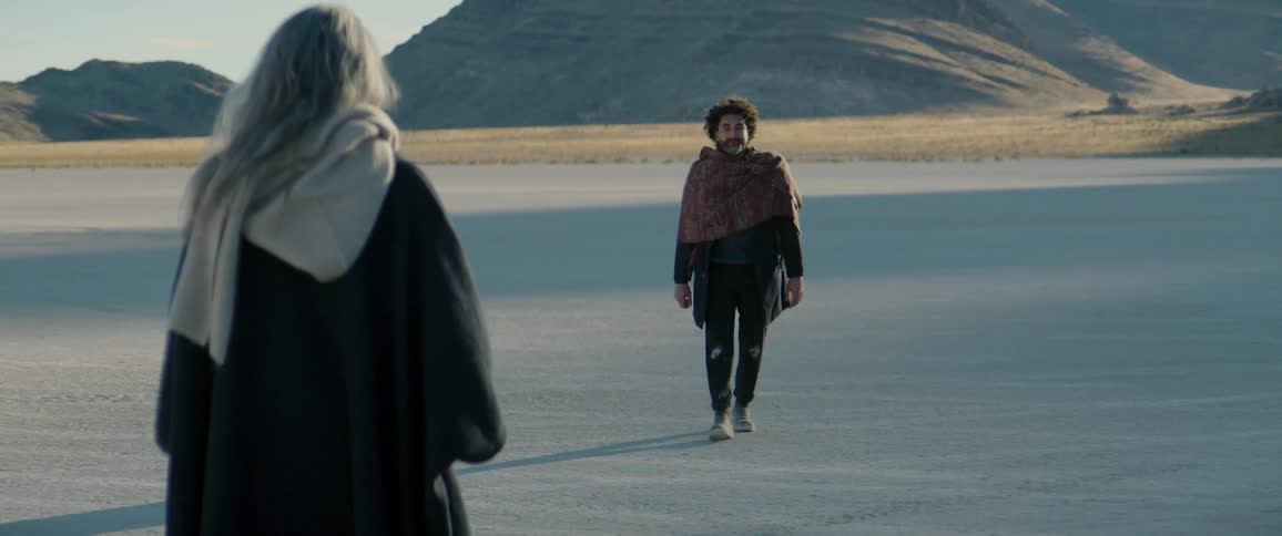 Man stands and walks across desert sands to meet approaching gray haired woman