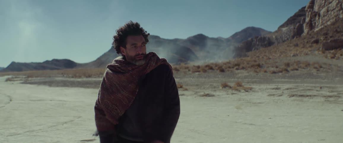 A man walking across a desert blows in to his hands to warm them as the wind blows