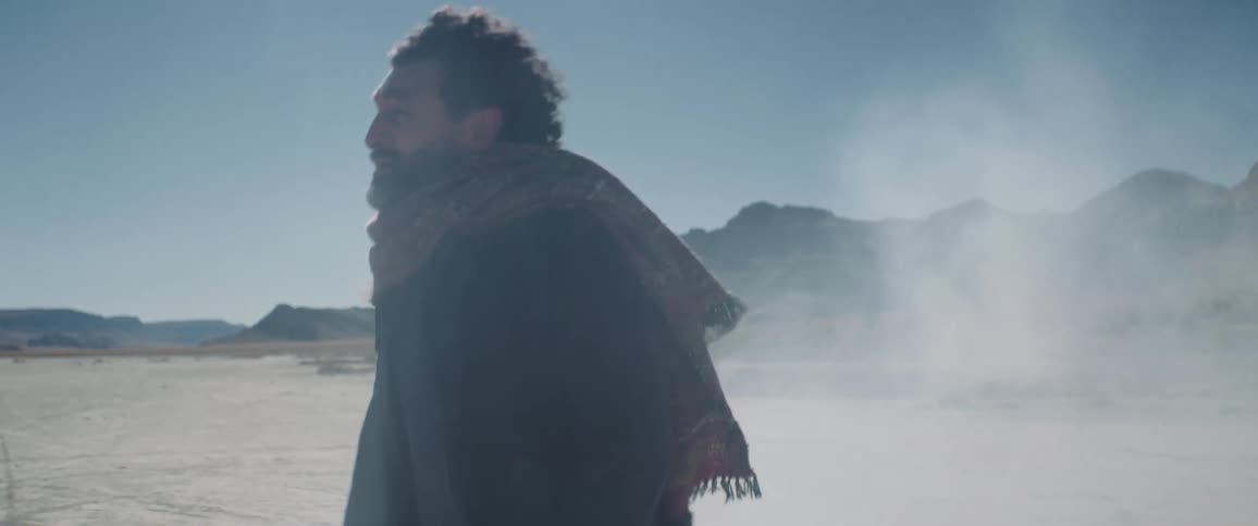 A man walking in a barren landscape shields his face from the wind while calling out for someone