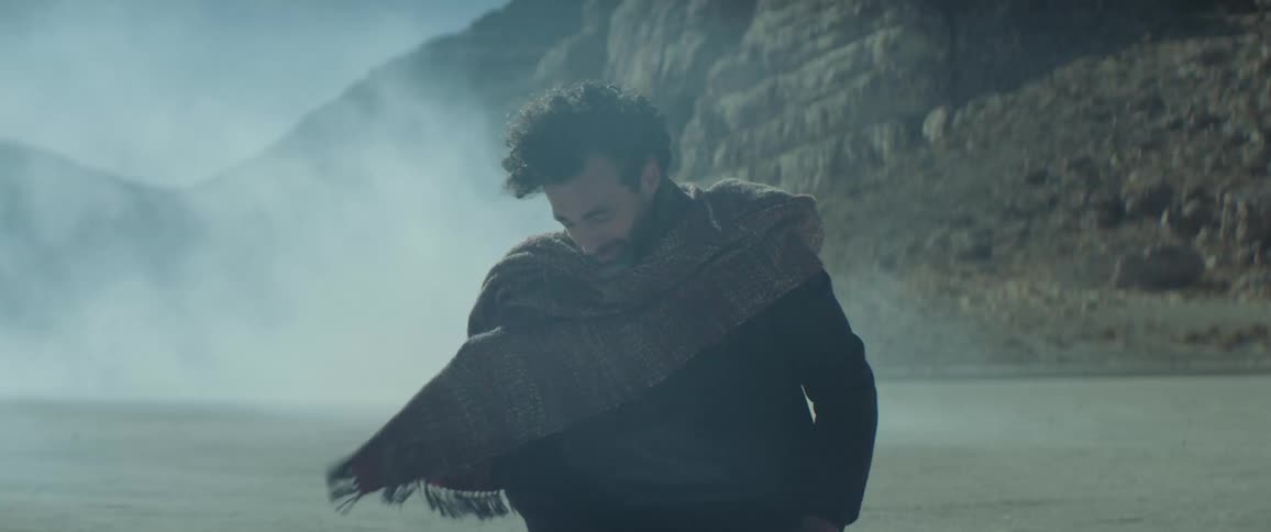 Man kneels on sand downcast as smoke smoke blows across area with mountain in the background