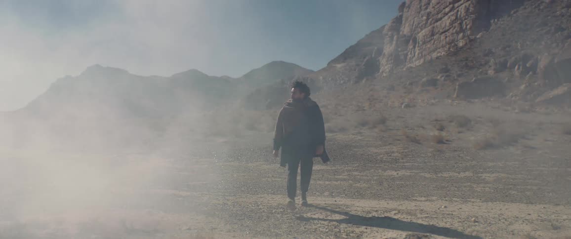 A man looks around at his surroundings as he walks through dust clouds in the badlands during the day