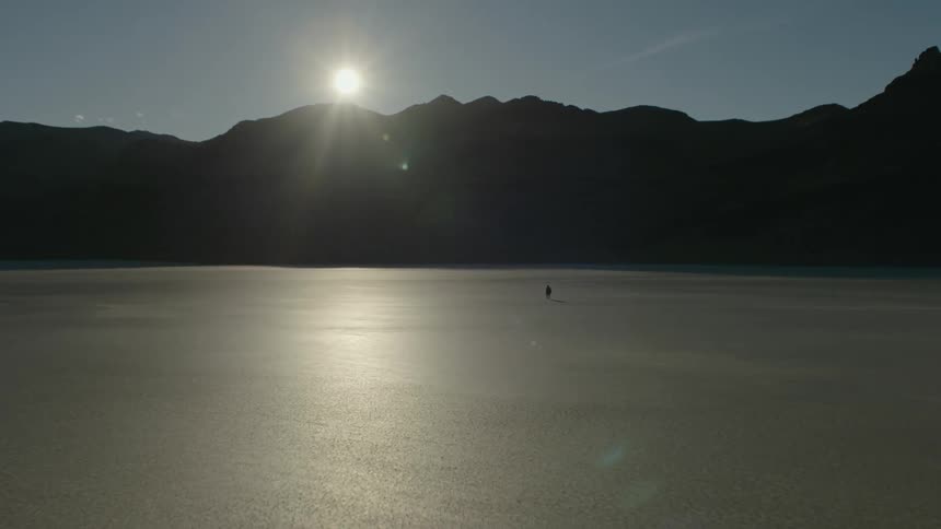 A person walks across a vast barren flatland towards a mountain ridge on a clear day
