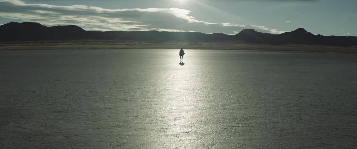 A silhouetted man walks across a barren flatland towards distant mountains during the day