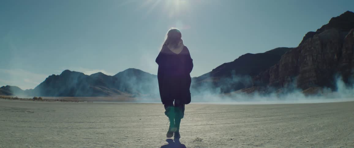 Gray haired woman walks across desert as smoke flows across ground near mountains on a sunny day