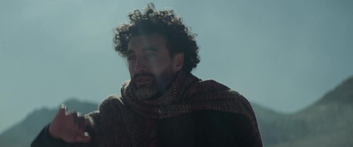 A man kneeling in a flatland raises his hand to shield his eyes as he looks into the distance before rising to his feet and beginning to walk