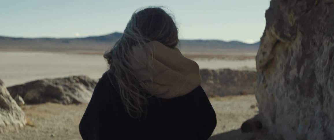 Gray haired person walks near stone wall facing mountain across a desert