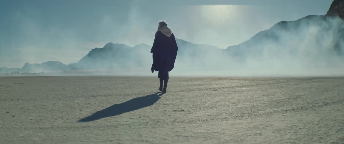 A women walking on cracked dry earth with fog and steam billowing past her as she looks around