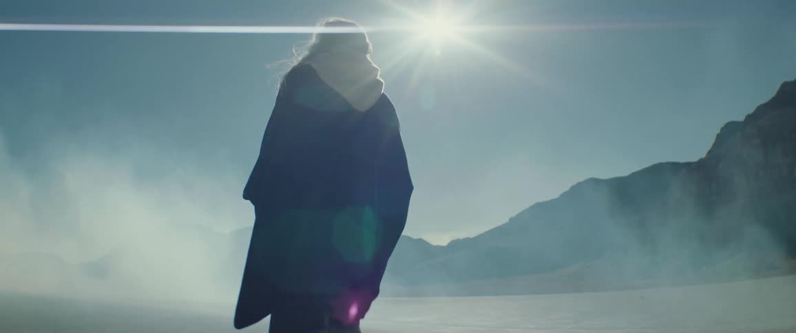 A woman walks through a barren flatland as clouds of dust float around her during the day