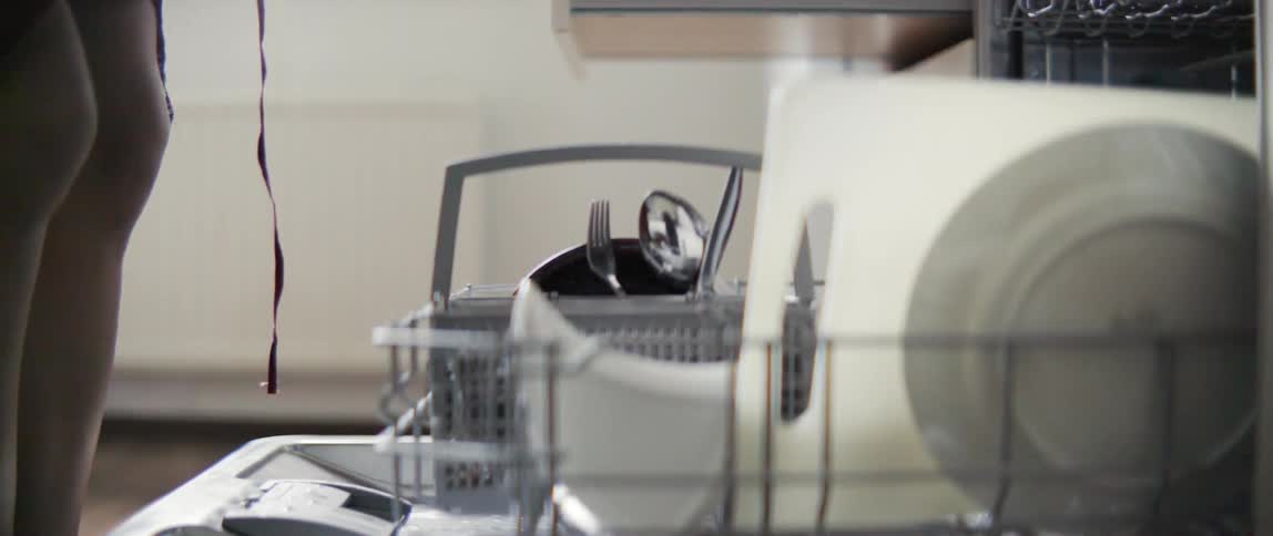 A woman carefully removes clean silver cutlery and utensils from a kitchen dishwasher rack.