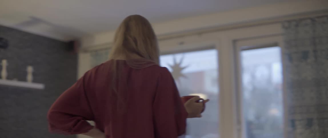 A woman walks through a messy living room while holding a warm coffee mug.