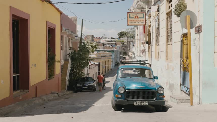 A blue vintage car parked on a town street in Cuba with pedestrians in the distance