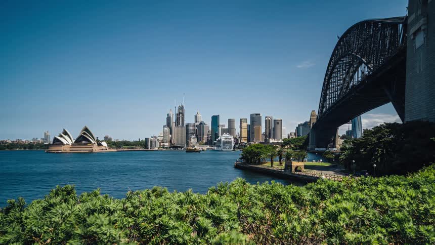 Boats in the harbor and people walking in the park near the sydney harbor bridge with the opera house and city skyline opposite