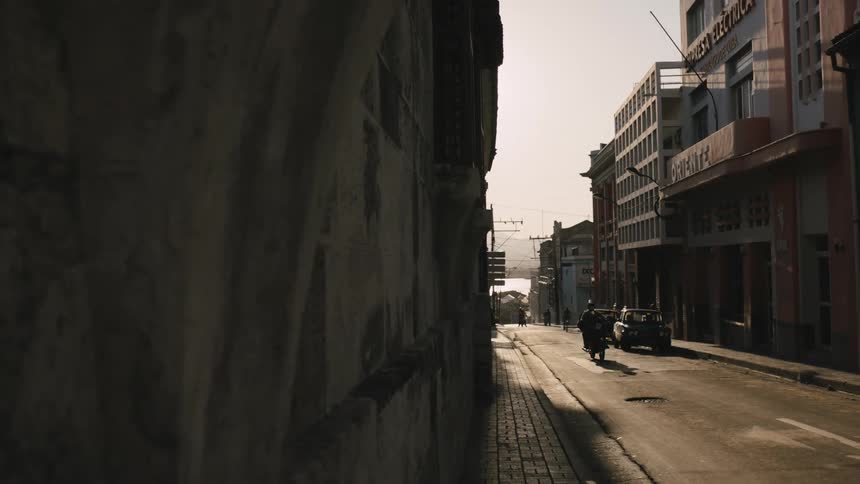 A motorcycle travels on a alley with cars parked on the roadside in front of buildings
