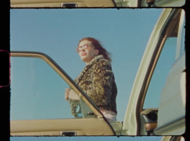 A woman adjusts her hair and clothing before leaning back to rest against a car outdoors on a clear day