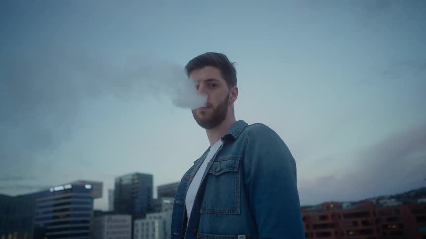 A man blows smoke out of his mouth while posing near downtown