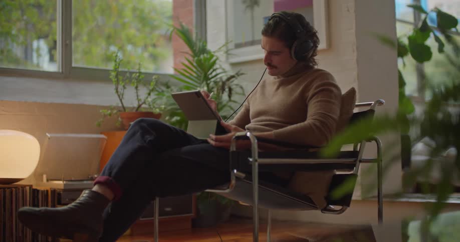A man sits at home and works on a tablet with headphones on