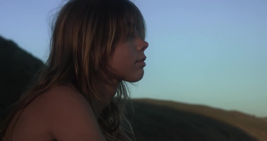 A teenage girl is sitting outside with mountains in the distance and the moon in a clear blue sky