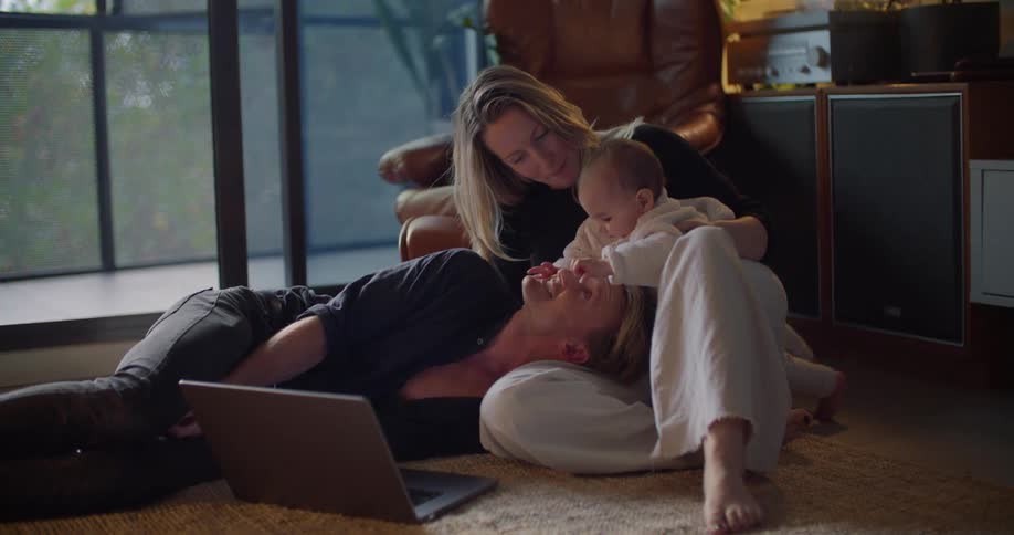 A man lies on the floor at home with his head in his wifes lap as their baby touches his face