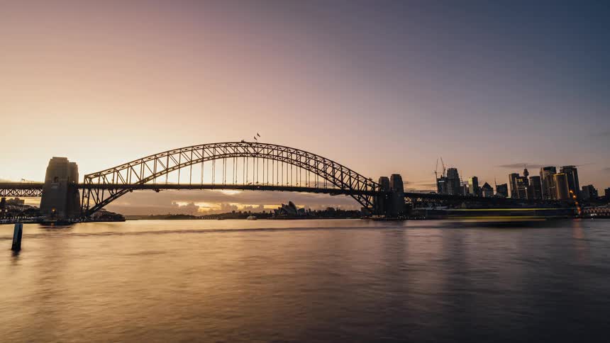 Boats and ships speed across water near bridge and city as night changes to early day