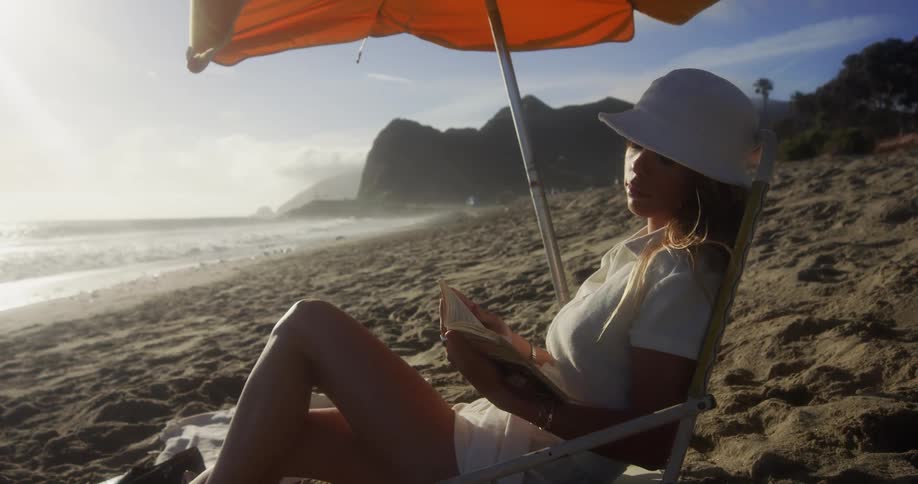 A woman wearing a sunhat is sitting in a chair on a beach reading a book near a sunshade umbrella