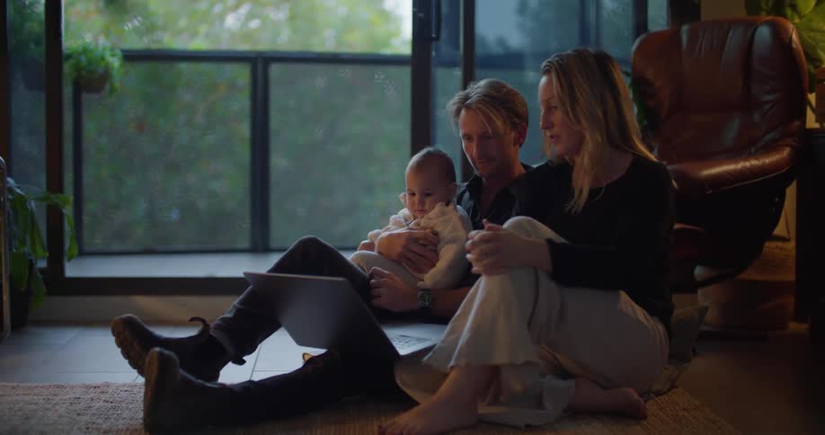 A man sitting on the living room floor with his baby in his lap talking with his wife pointing at a laptop