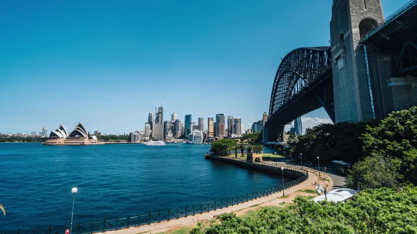 Pedestrians and traffic move along shore near bridge over water across from sydney opera house and downtown