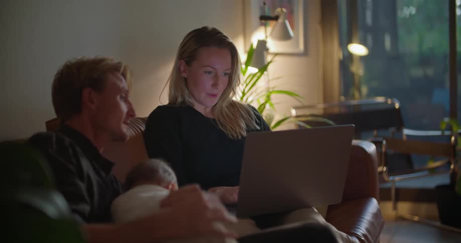 Mother and father sit on couch with baby discussing contents of laptop screen as mother types