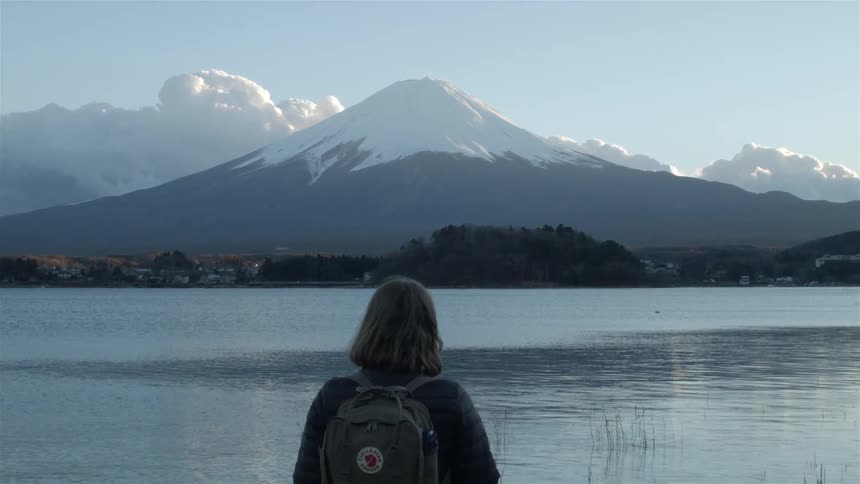 A young woman carrying a backpack stares at a snow capped mountain above a serene lake