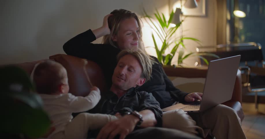 Baby climbs over reclining father as mother uses laptop