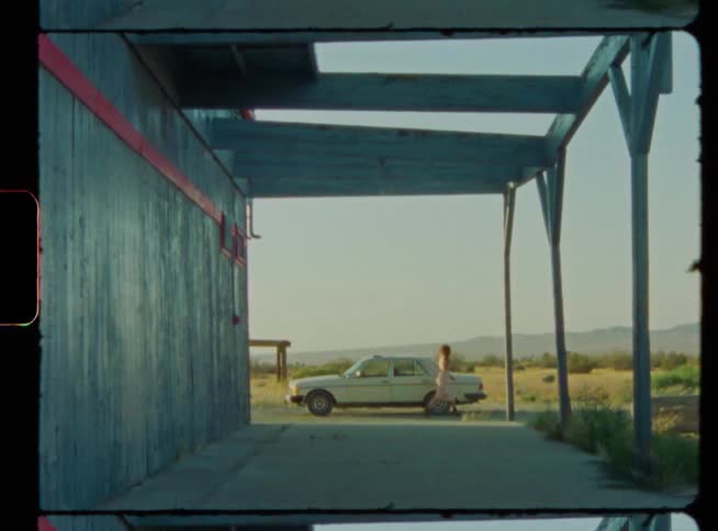 A car is parked near a building in a rural area and a woman gets out and walks to the trunk