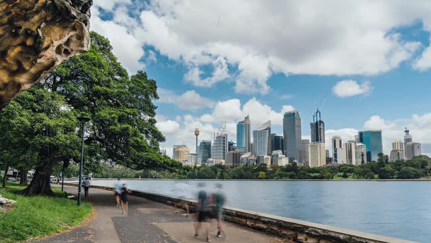 Pedestrians and joggers move along path through park across water from city skyline