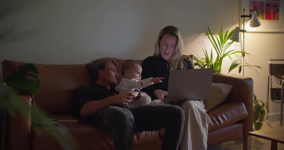 A woman is sitting on the couch typing on a laptop computer as her husband sits next to her holding their baby