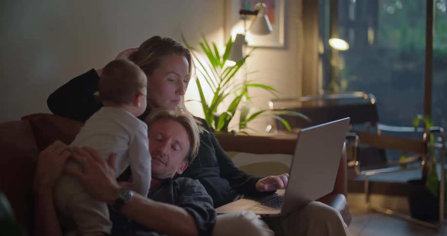 Family occupies couch the parents interacting with baby as mother types on laptop