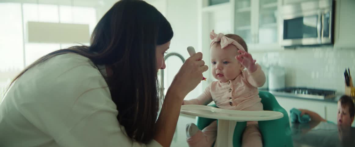 A woman feeds her baby at home while her young son wipes a counter
