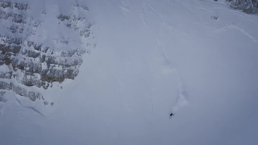 Snowboarder and others wait on slope as skier skis down snow covered mountain side spraying snow and leaving tracks