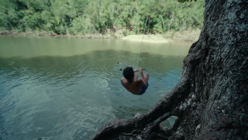 A boy holds onto a rope swing hanging from a tree then swings out and splashes into a pond