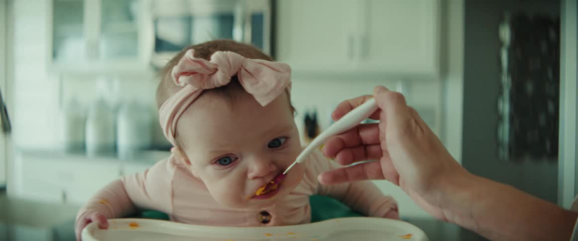 A woman feeds her baby while the girl sits in a high chair in the kitchen