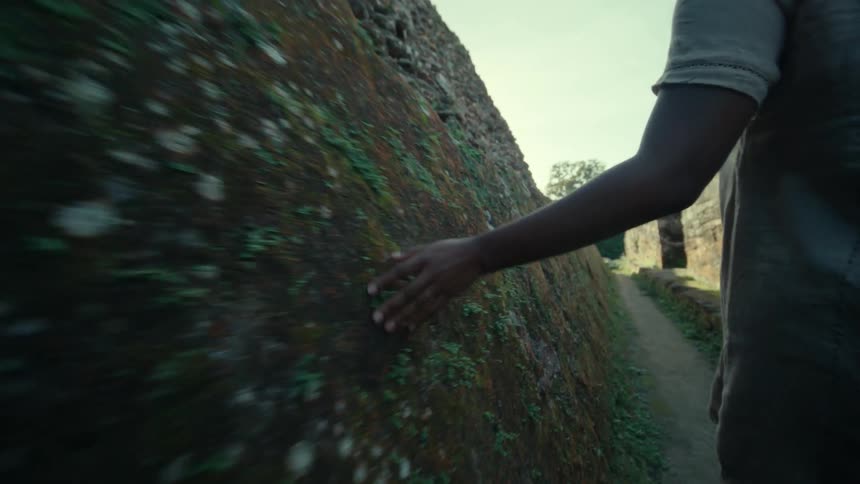 Woman walks along path through ruins touching rough rock walls