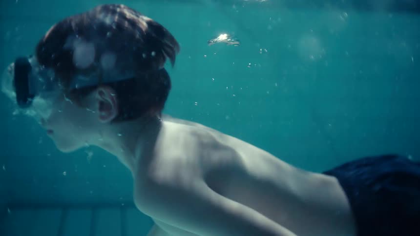 An underwater view of boy in a swimming pool swimming to the bottom and back to the surface while wearing goggles