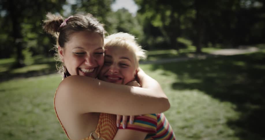 Woman holds up sign with the words free hugs then hugs a female friend as they stand in the park