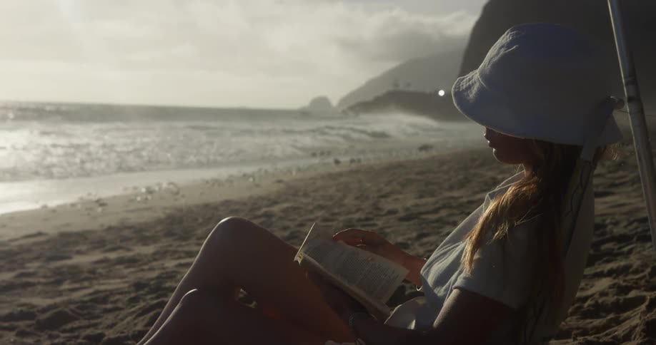 A woman wearing a sunhat is sitting on a beach reading a book