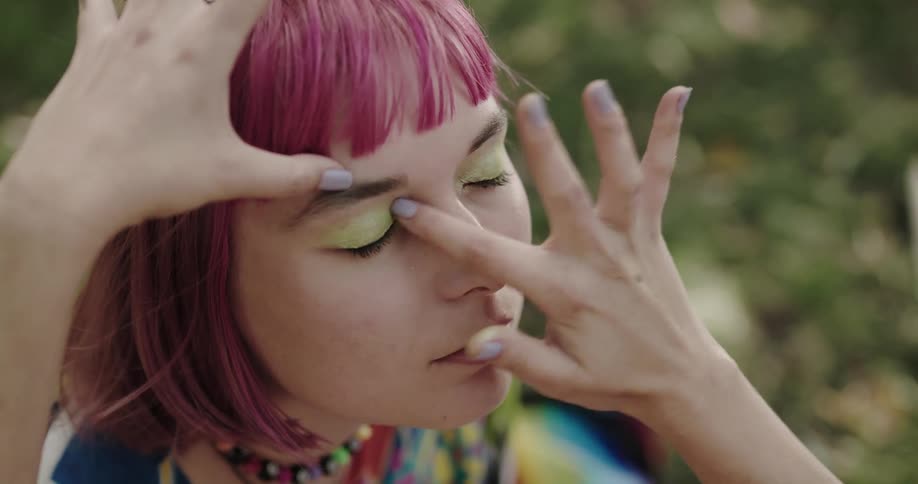 A teenage girl with pink hair is sitting outside and she closes her eyes as another girl fixes her eye makeup