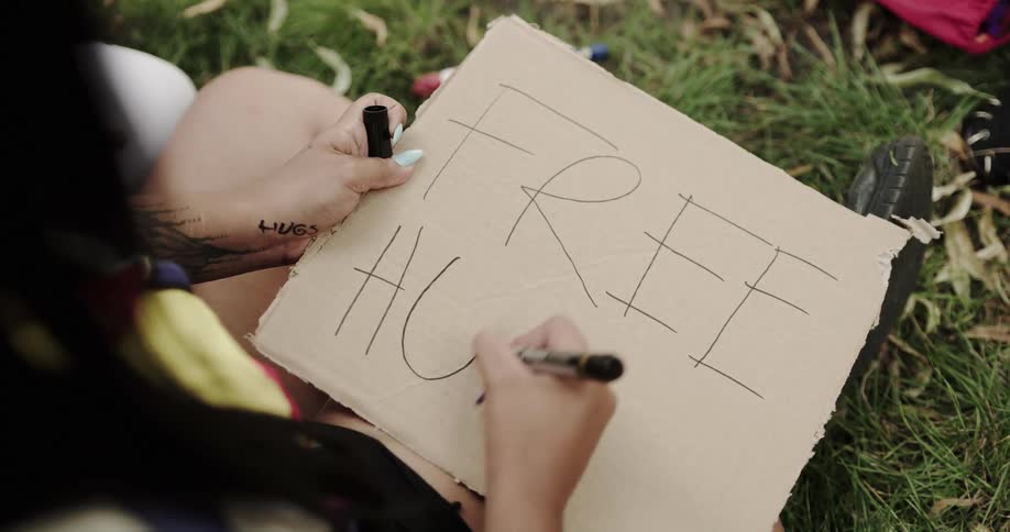 Woman sitting on grass writes sign about free hugs