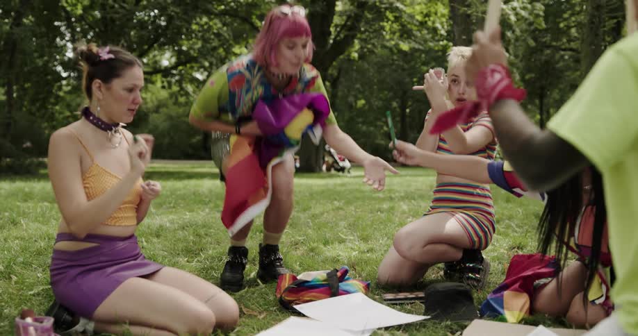 A group of friends cheer as a woman stands up holding a rainbow pride flag in a park