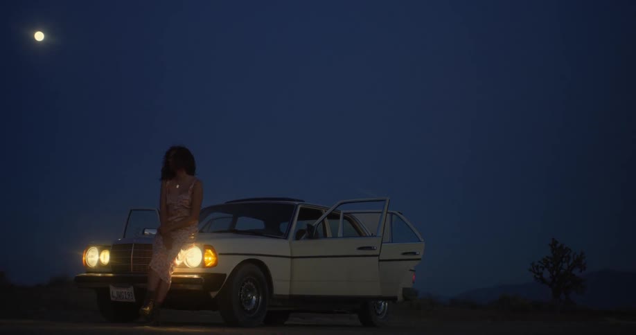 A woman leans against the front of her car at night