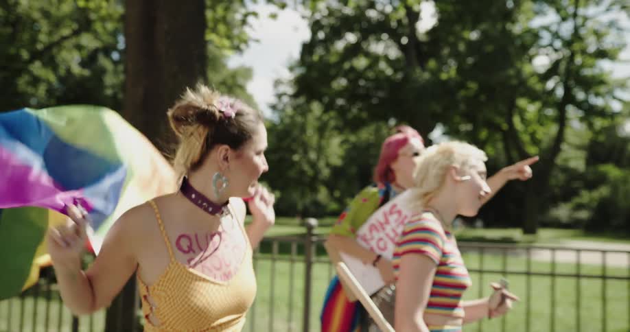 Teenagers walk down a street past a public park carrying signs and waving a rainbow flag