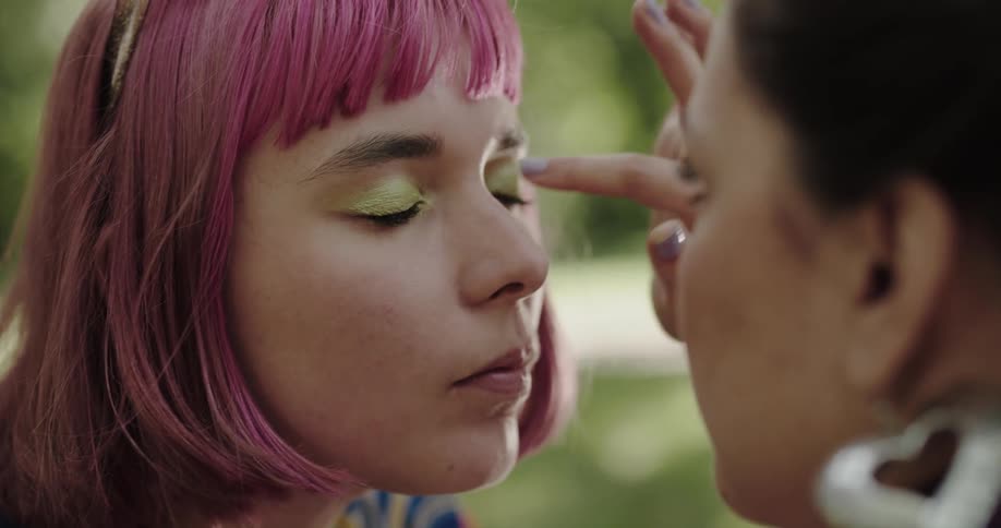 A teenage girl with pink hair is sitting outside and she closes her eyes as another girl fixes her eye makeup