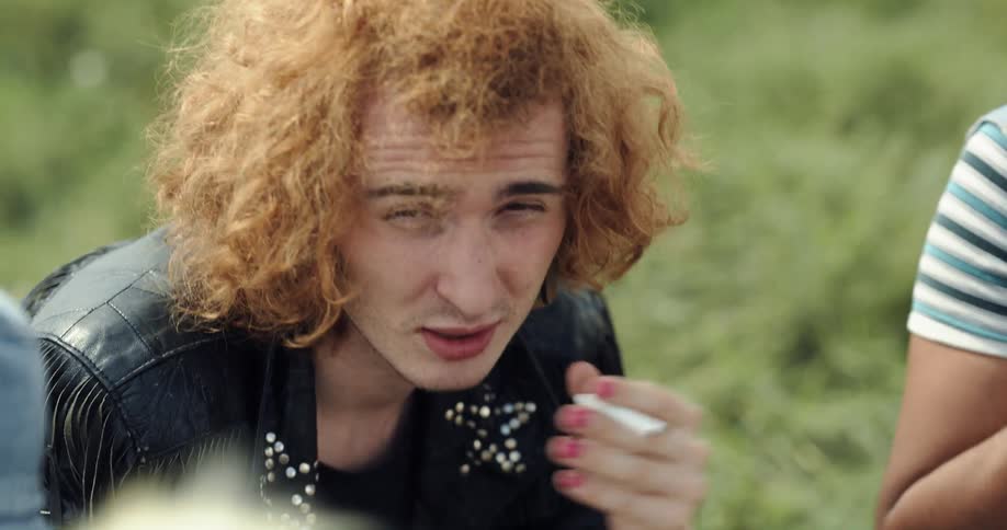 A man places a cigarette in his mouth and prepares to light it while chatting with a group of people sitting with him in a field during the day