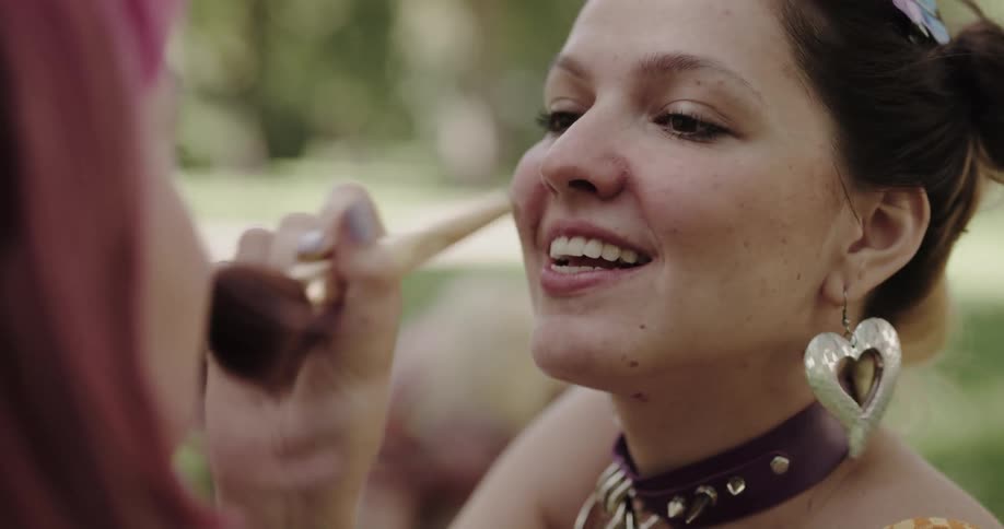 A smiling makeup artist applying cosmetics to a woman with purple hair outside