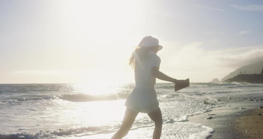 A woman holds her sunhat as she run across a beach as the waves roll in
